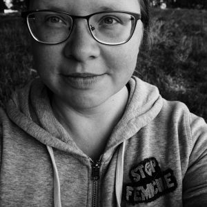 von der Autorin gemachtes Selbstporträt in schwarz-weiss, sie trägt eine große Brille und auf ihrem Kapuzenpullover ist ein Aufnäher gestickt: Stop Femicide. Sie schaut links an der Kamera vorbei und lächelt etwas.